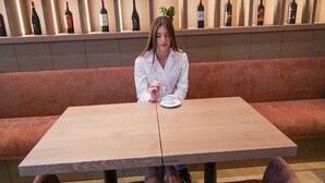 A person seated at a table with a white tablecloth, surrounded by a red cushioned bench and two red upholstered chairs. Behind them is a wall with shelves holding various bottles and potted plants. - Post Inn  (Tirana)