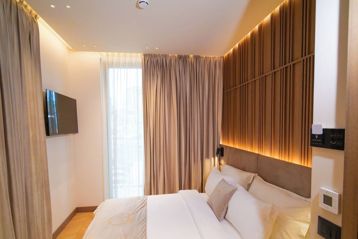A modern hotel room with a large bed, a flat-screen TV mounted on the wall, and a control panel for room amenities.