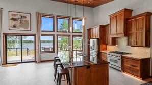 A modern kitchen with wooden cabinets, a large island with bar stools, and a view of a lake through large windows. - Escape to Lakeview Paradise, a luxury 4-bed, 3-bath lakefront retreat in Tripoli, WI. Each bedroom has a king bed, and the 2,560 sq. ft. home features high-end finishes, cozy comforts, and stunning lake views from a private 24-ft deck or hot tub. (Tripoli)