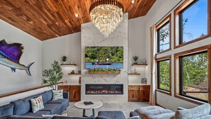 A modern living room with a fireplace, a mounted TV, and a large window overlooking a forest. - Escape to Lakeview Paradise, a luxury 4-bed, 3-bath lakefront retreat in Tripoli, WI. Each bedroom has a king bed, and the 2,560 sq. ft. home features high-end finishes, cozy comforts, and stunning lake views from a private 24-ft deck or hot tub. (Tripoli)