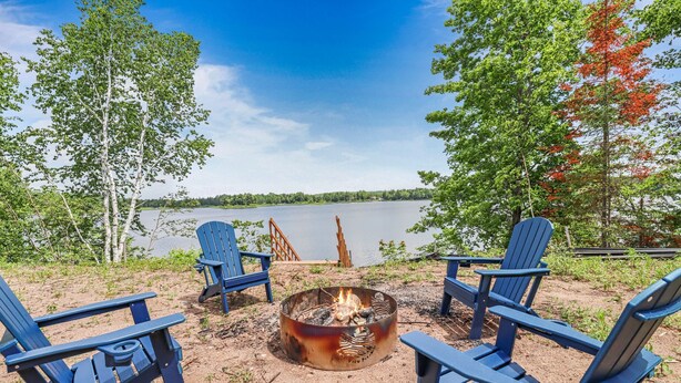 A lakeside setting with a fire pit surrounded by Adirondack chairs, overlooking a calm lake and forested area. - Escape to Lakeview Paradise, a luxury 4-bed, 3-bath lakefront retreat in Tripoli, WI. Each bedroom has a king bed, and the 2,560 sq. ft. home features high-end finishes, cozy comforts, and stunning lake views from a private 24-ft deck or hot tub. (Tripoli)