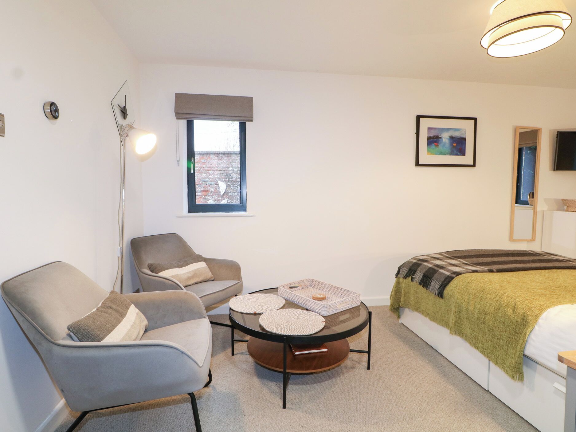 Une chambre moderne avec un lit, deux chaises, une petite table, une lampe sur pied et une fenêtre donnant sur un mur en briques.