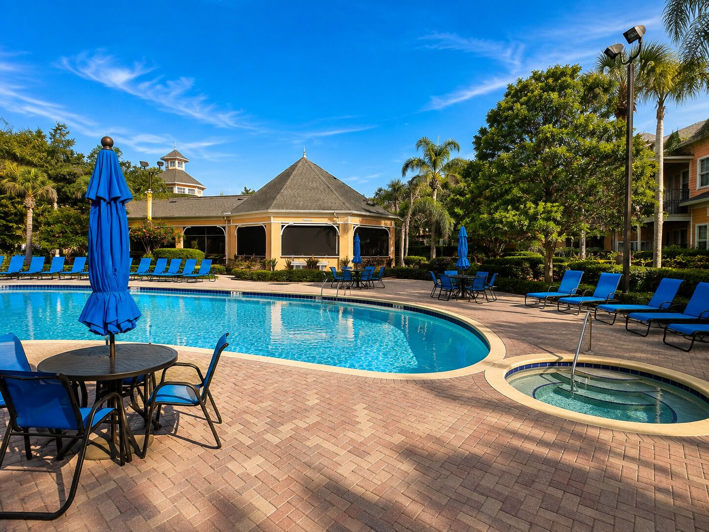 A swimming pool with lounge chairs and umbrellas, surrounded by a brick patio and palm trees.