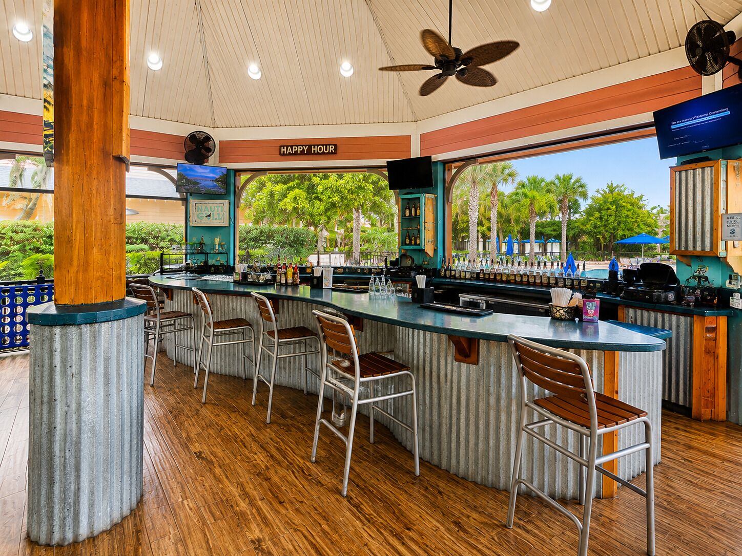 A bar area with a wooden floor, metal bar stools, a counter with bottles, and a ceiling fan.