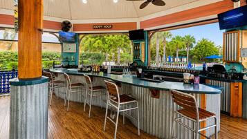 A bar area with a wooden floor, metal bar stools, a counter with bottles, and a ceiling fan.