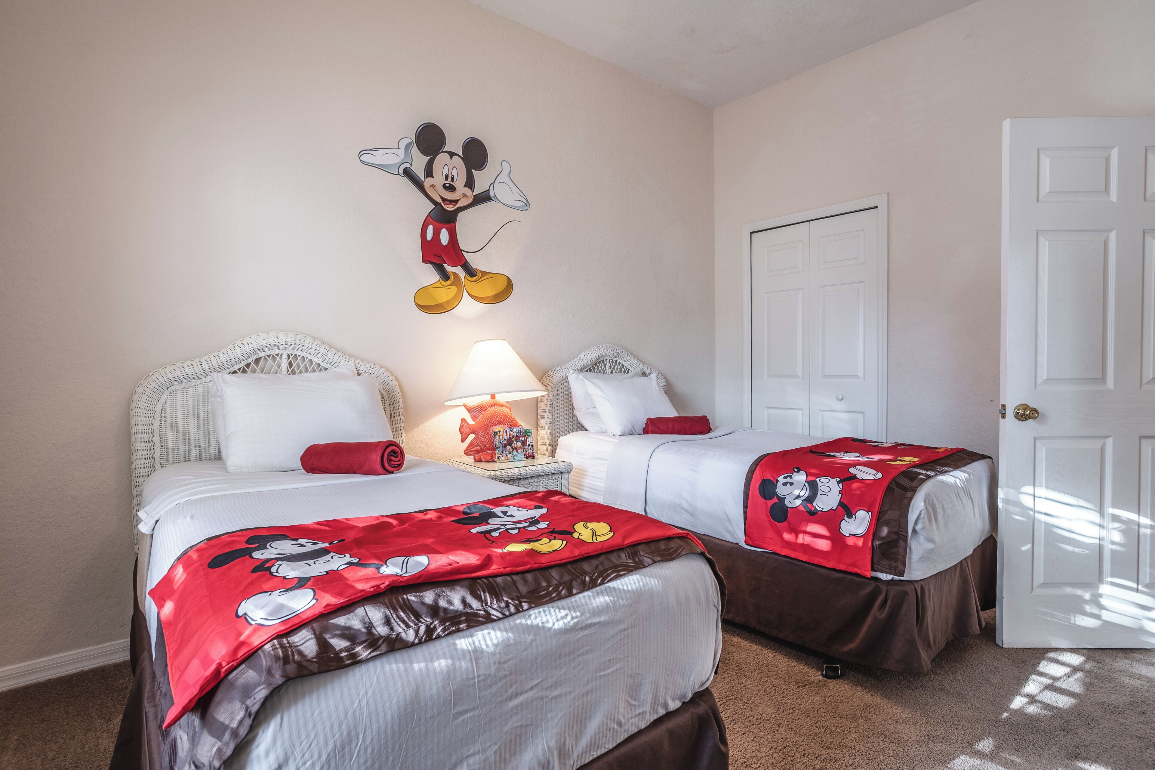 A hotel room with two beds, each with Mickey Mouse-themed bedding. There is a wall-mounted Mickey Mouse decoration and a lamp on a nightstand.