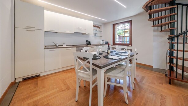 A modern kitchen with white cabinets, a dining table set for four, and a spiral staircase. - ApartmentsArena - Capitolium Verona 6 (Verona)
