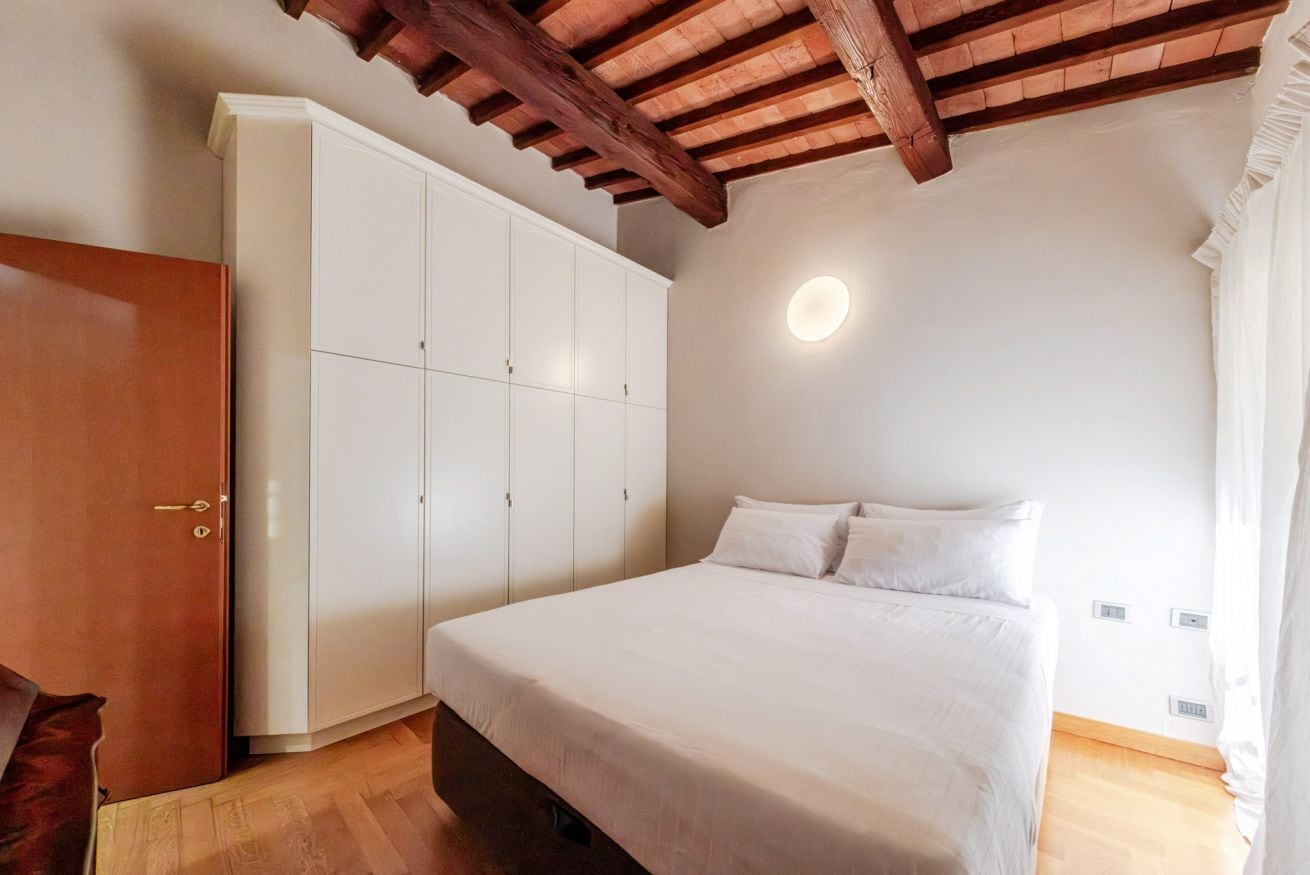 Ein Schlafzimmer mit Bett, weißem Wandschrank, Holzbalken an der Decke und einer runden Deckenleuchte.
