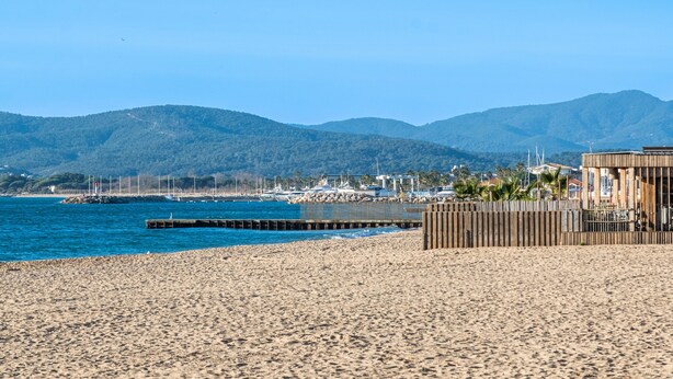 A beach with a wooden structure, a pier, and mountains in the background. - Doux Zéphir – Marine Serenity and Tranquility by the Sea (Fréjus)
