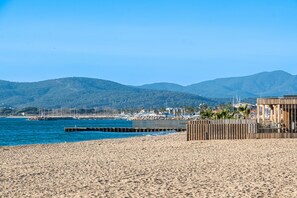 A beach with a wooden structure, a pier, and mountains in the background. - Doux Zéphir – Marine Serenity and Tranquility by the Sea (Fréjus)