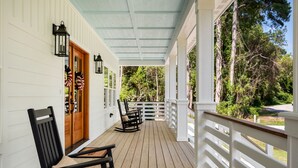 A wooden deck with a white railing, a rocking chair, and a door with a wreath. - Discover this stunning award-winning luxury 4br & 3.5 bath home in the OBX (Powells Point)