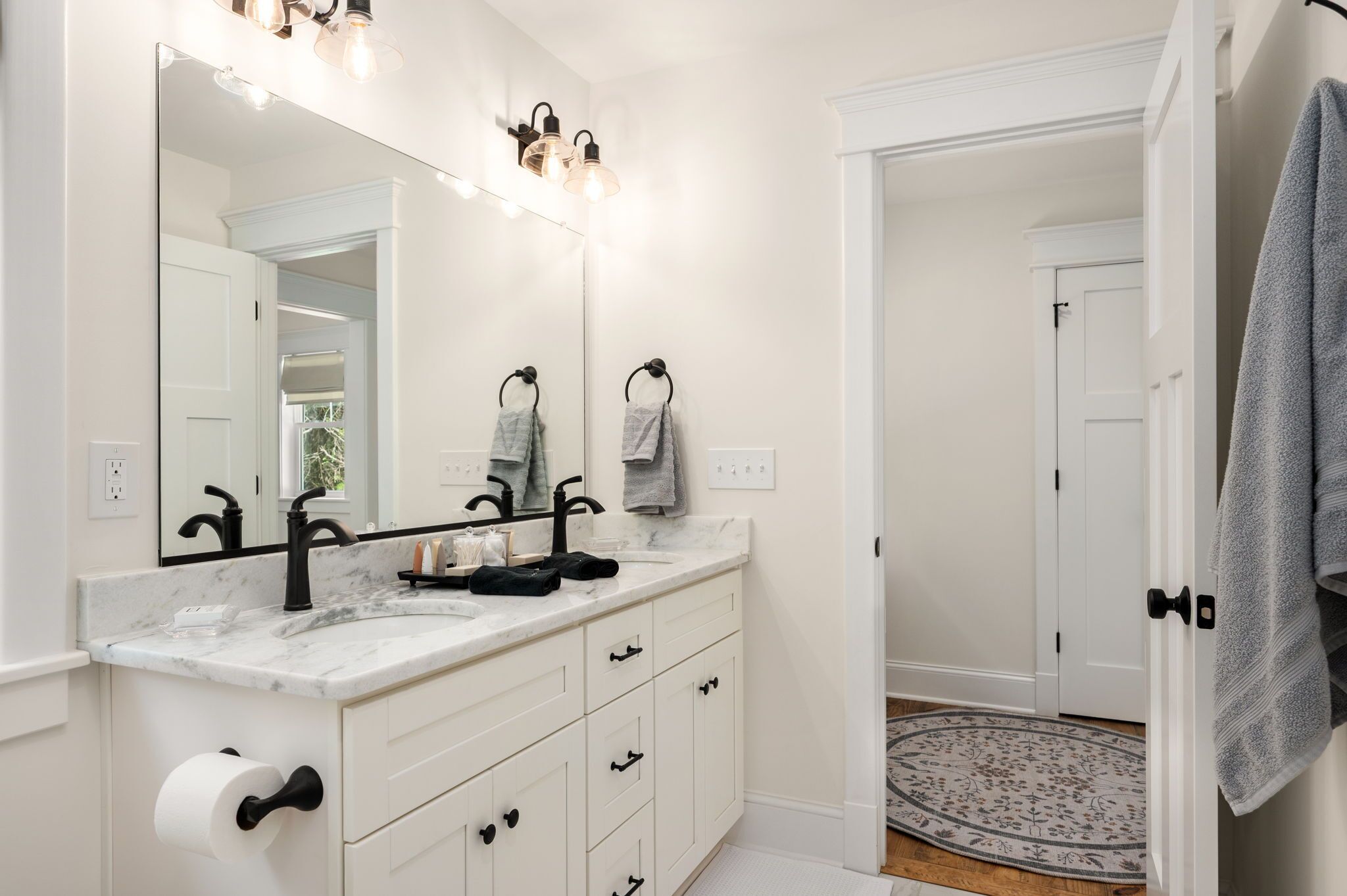 Une salle de bain avec un meuble-lavabo blanc, des robinets noirs, un grand miroir avec éclairage intégré et un porte-serviettes.