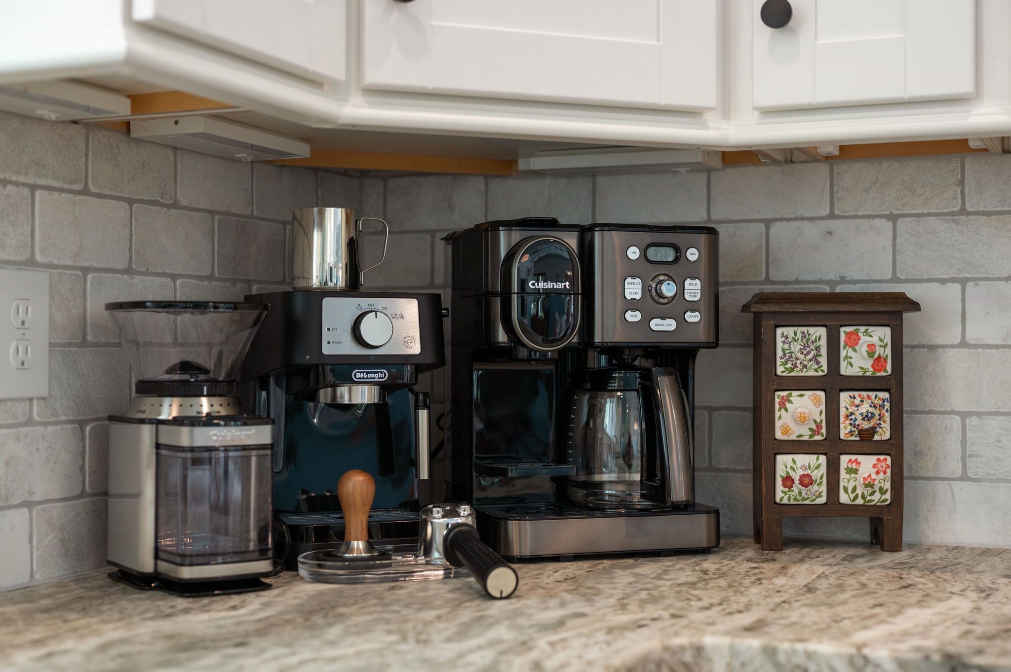 A kitchen with a coffee maker, a French press, and a small cabinet with floral tiles.