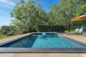 Une piscine rectangulaire entourée d’une terrasse en bois, une pelouse avec des chaises longues et une haie en arrière-plan.