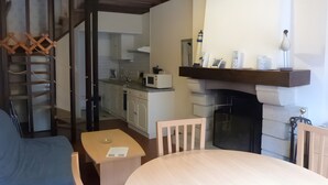 Een kamer met een eettafel, stoelen, een keukenhoek met kasten en apparatuur, een open haard en een trap.