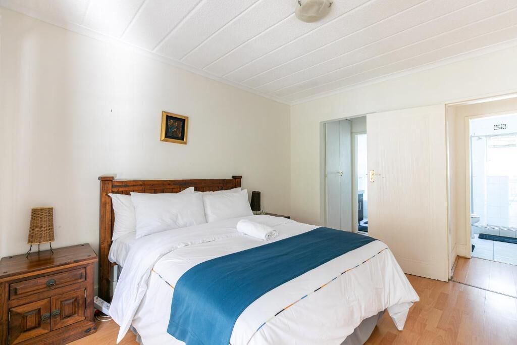 Une chambre à coucher avec un lit en bois, une literie blanche, une touche de couleur bleue, une table de chevet et une porte donnant sur une salle de bain.