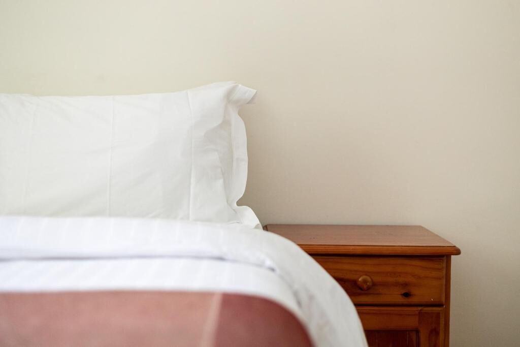 Una cama con ropa de cama blanca y una mesita de noche de madera con cajón.