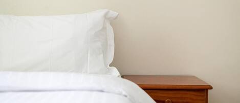 Una cama con ropa de cama blanca y una mesita de noche de madera con cajón.