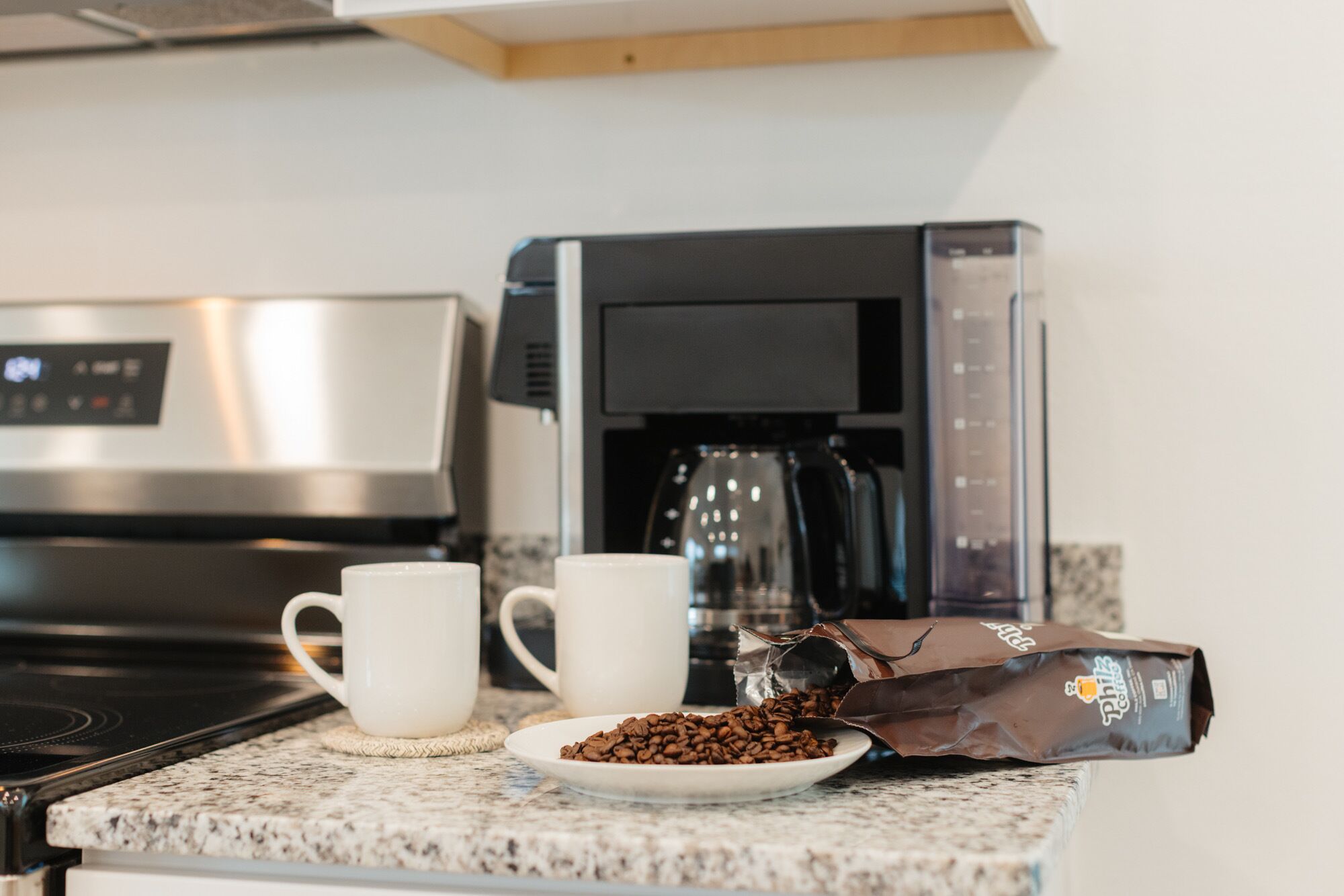 A kitchen counter with a coffee maker, two white cups, a plate of coffee beans, and a coffee bag.