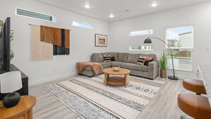 A modern living room with a sectional sofa, a coffee table, and a large window providing natural light. - Modern 3BR Centerton Near Walmart HQ & Bentonville (Centerton)