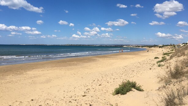 Ein weiter Sandstrand mit sanften Wellen, blauem Himmel und vereinzelten Wolken.