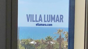Blick aus einem Fenster der Villa Lumar, der eine Küstenlandschaft mit Palmen und einem klaren Himmel zeigt.