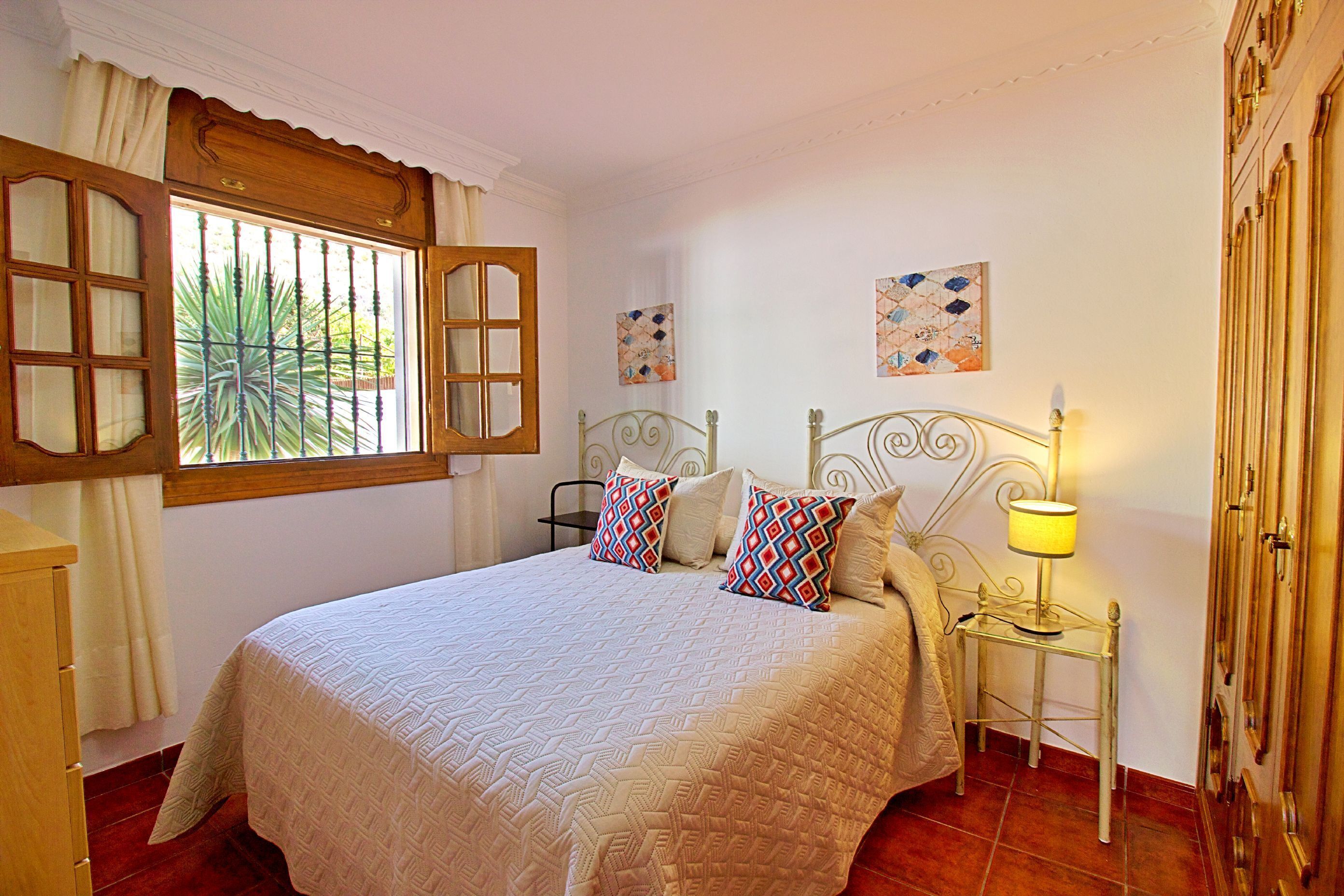 A neatly made bed with a patterned bedspread and decorative pillows, a bedside table with a lamp, and a window with curtains.