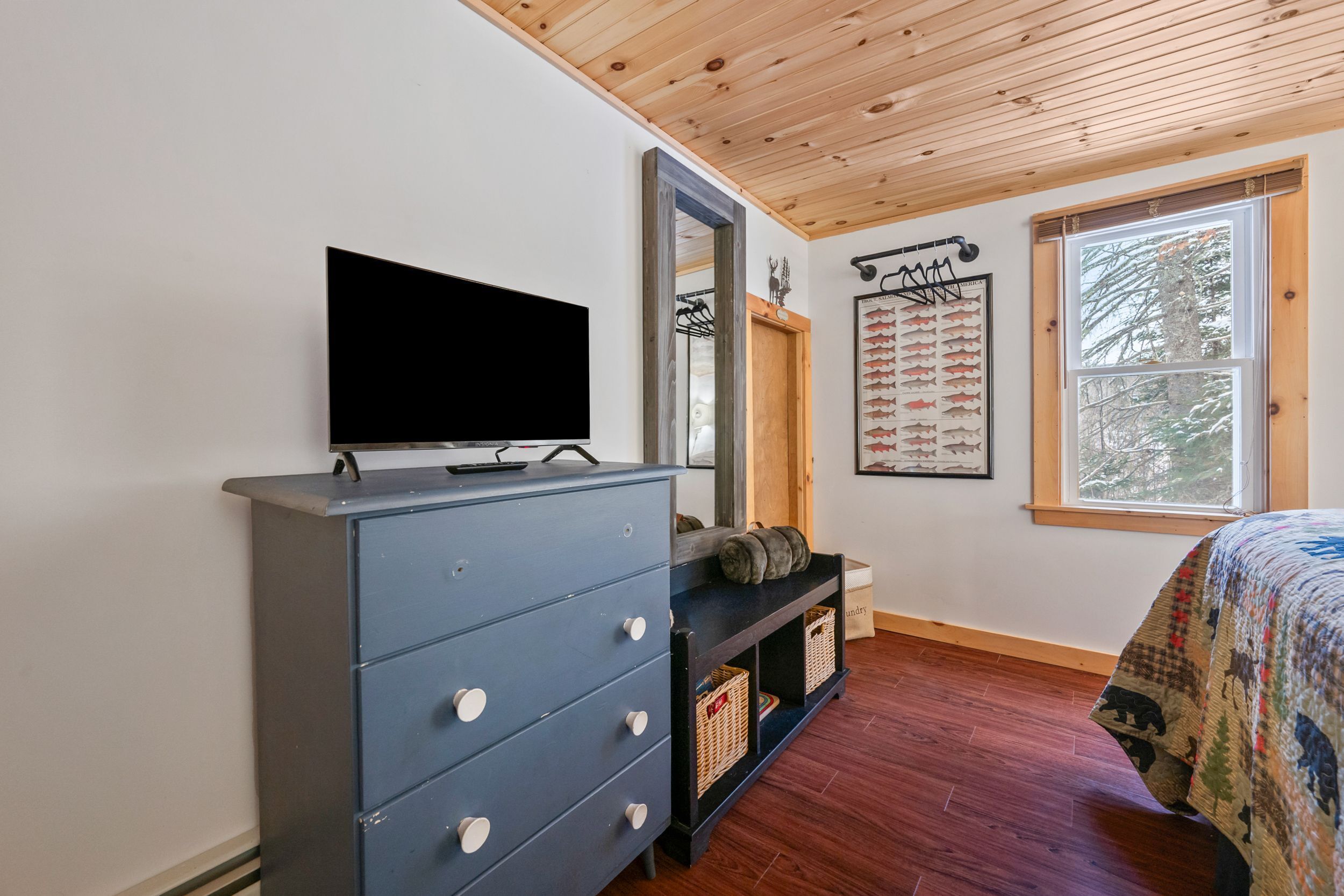 A bedroom with a wooden ceiling, a dresser with drawers, a television on top, a bed with a patterned blanket, a window with a view of trees, and a wall-mounted coat rack.