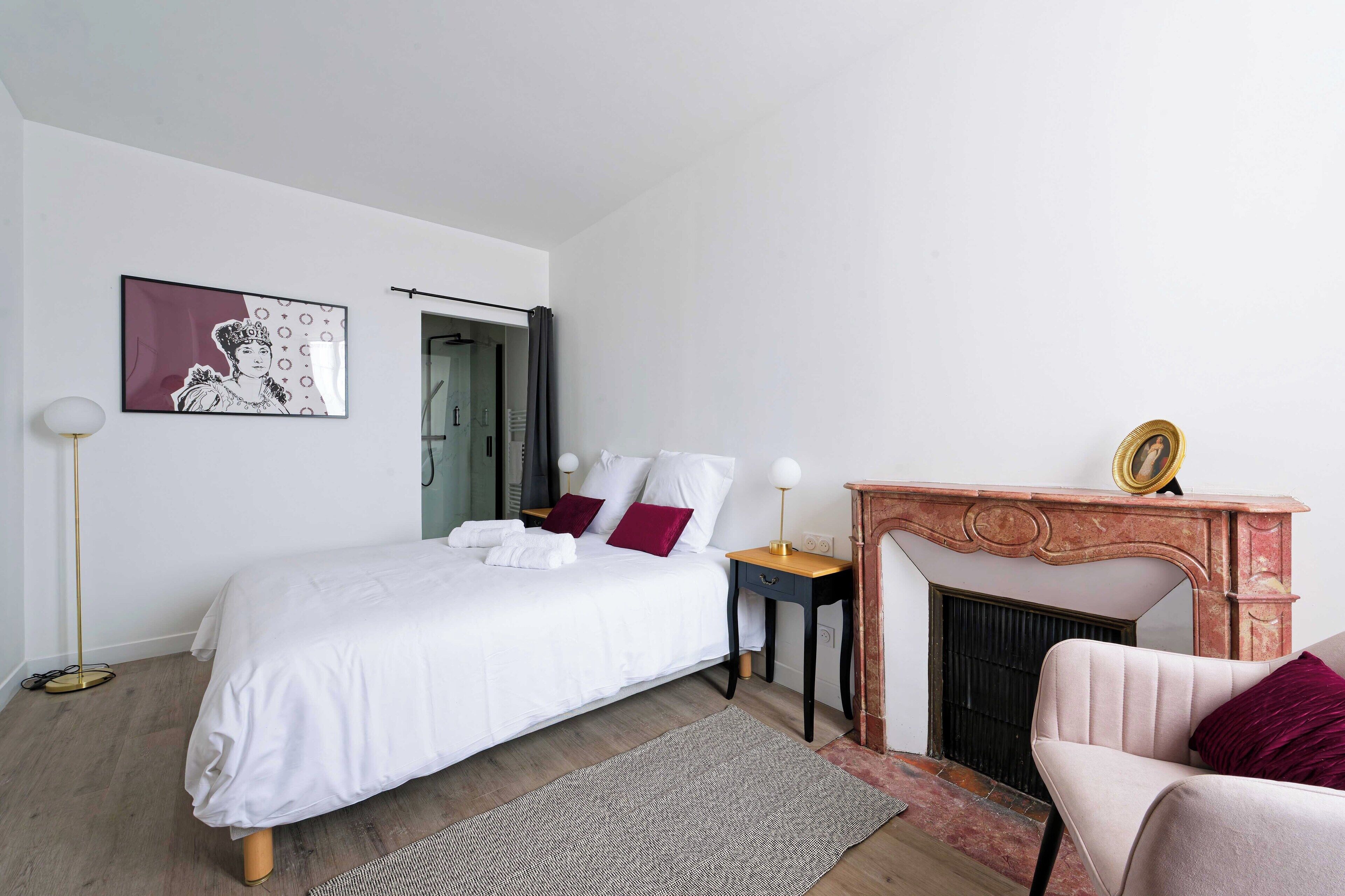 A bedroom with a bed, a nightstand, a chair, a fireplace, and a picture on the wall.
