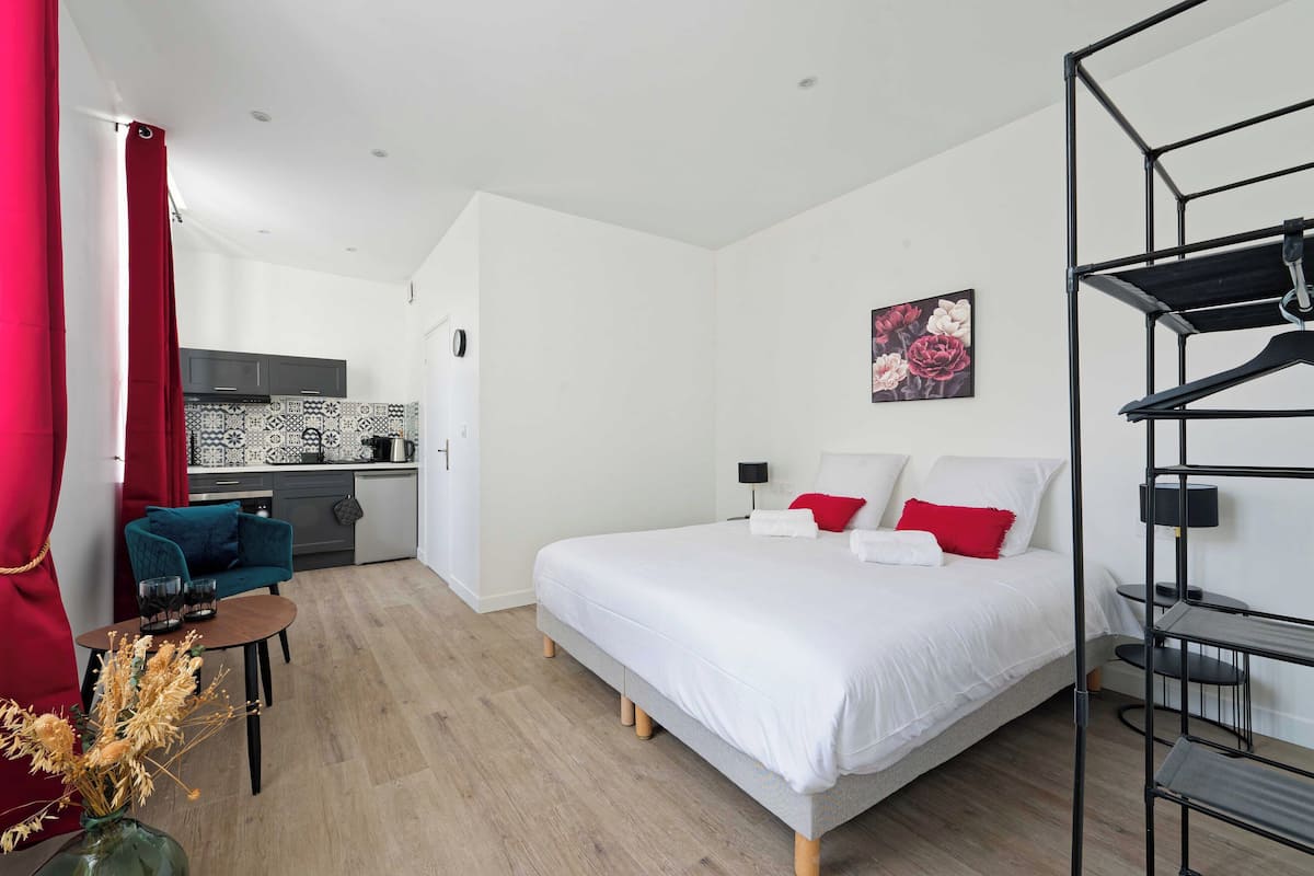 A modern hotel room with a bed, a small table with a vase, a kitchenette, and a red curtain.