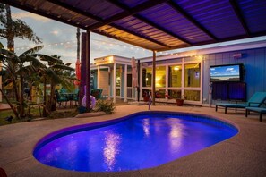 Una piscina con un porche cubierto, mobiliario de exterior y vistas a un edificio con grandes ventanales.