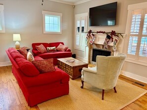 A living room with a red sofa, a white armchair, a wooden coffee table, and a fireplace with holiday decorations. - Charming Walkable Downtown Southern Pines 3 bed/2-1/2 Bath! (Southern Pines)
