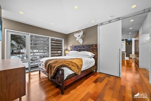 Une chambre moderne avec un lit en bois, une grande fenêtre avec des stores et une sculpture représentant une tête de cerf fixée au mur.