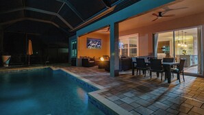 An indoor swimming pool area with a patio and seating, a ceiling fan, and a television screen displaying a sports event. - Coral Oasis: Screened Lanai with Private Heated Pool & Spacious 4-Bed Retreat (Cape Coral)