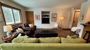 En stue med grøn sofa, sort fodskammel, træsofa bord, gulvlampe og bogreol.