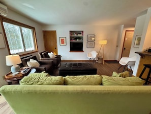 A living room with a green sofa, a black ottoman, a wooden coffee table, a floor lamp, and a bookshelf.