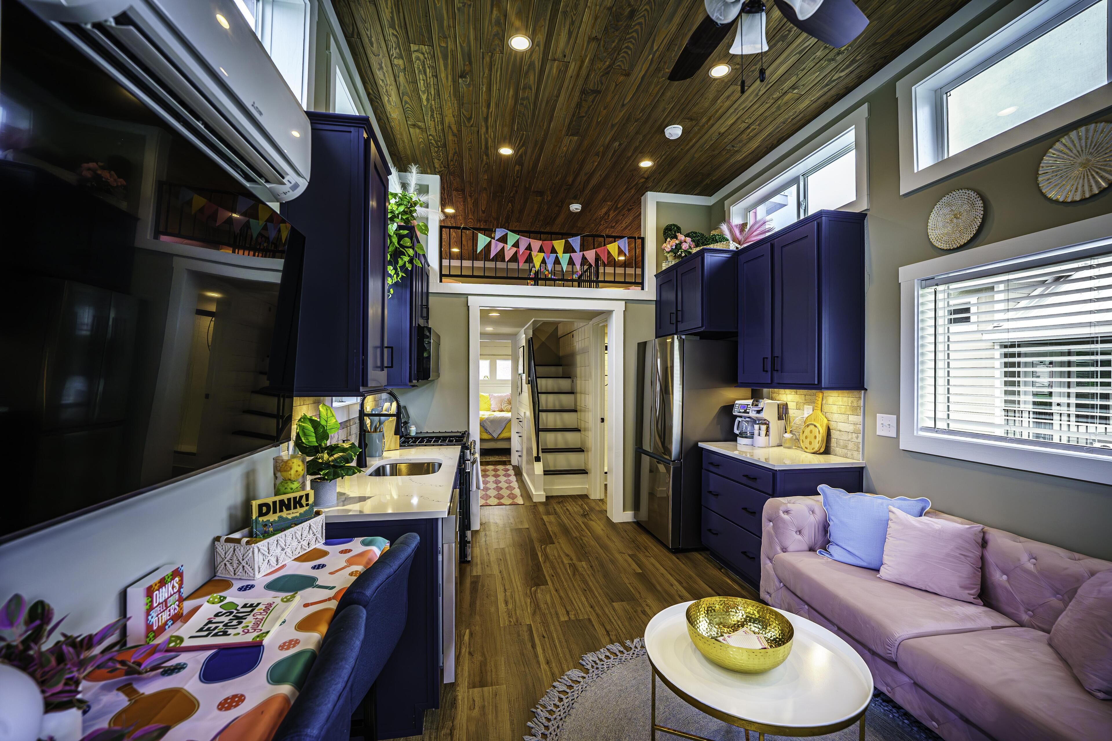 Een compacte keuken met een centraal eiland, een eethoek met een tafel en stoelen, een roze bank en een trap naar een verdieping erboven.