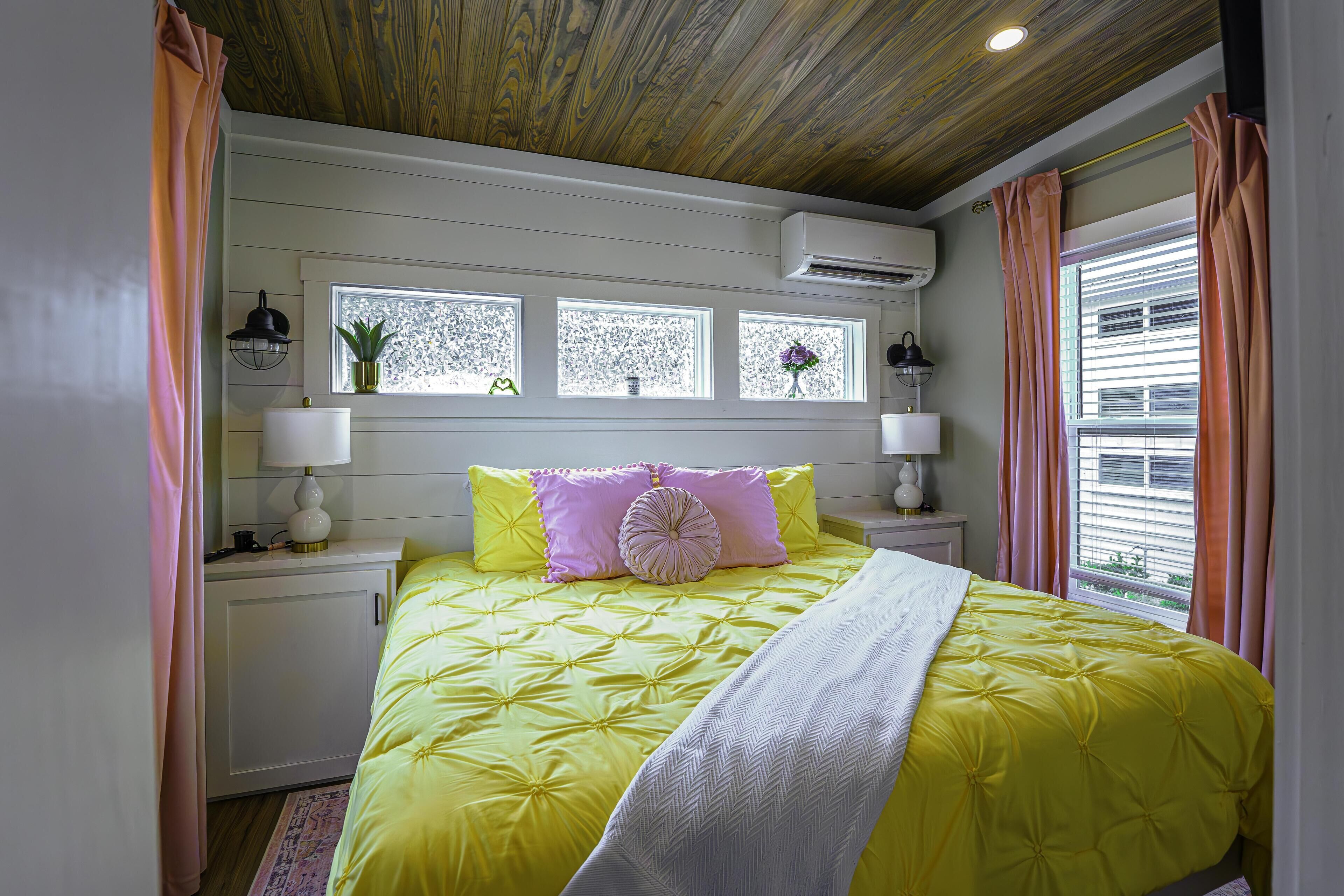Une chambre avec une couvre-lit jaune, une tête de lit blanche et des rideaux roses. Il y a trois fenêtres ornées de grilles décoratives, et un appareil de climatisation blanc placé au-dessus.