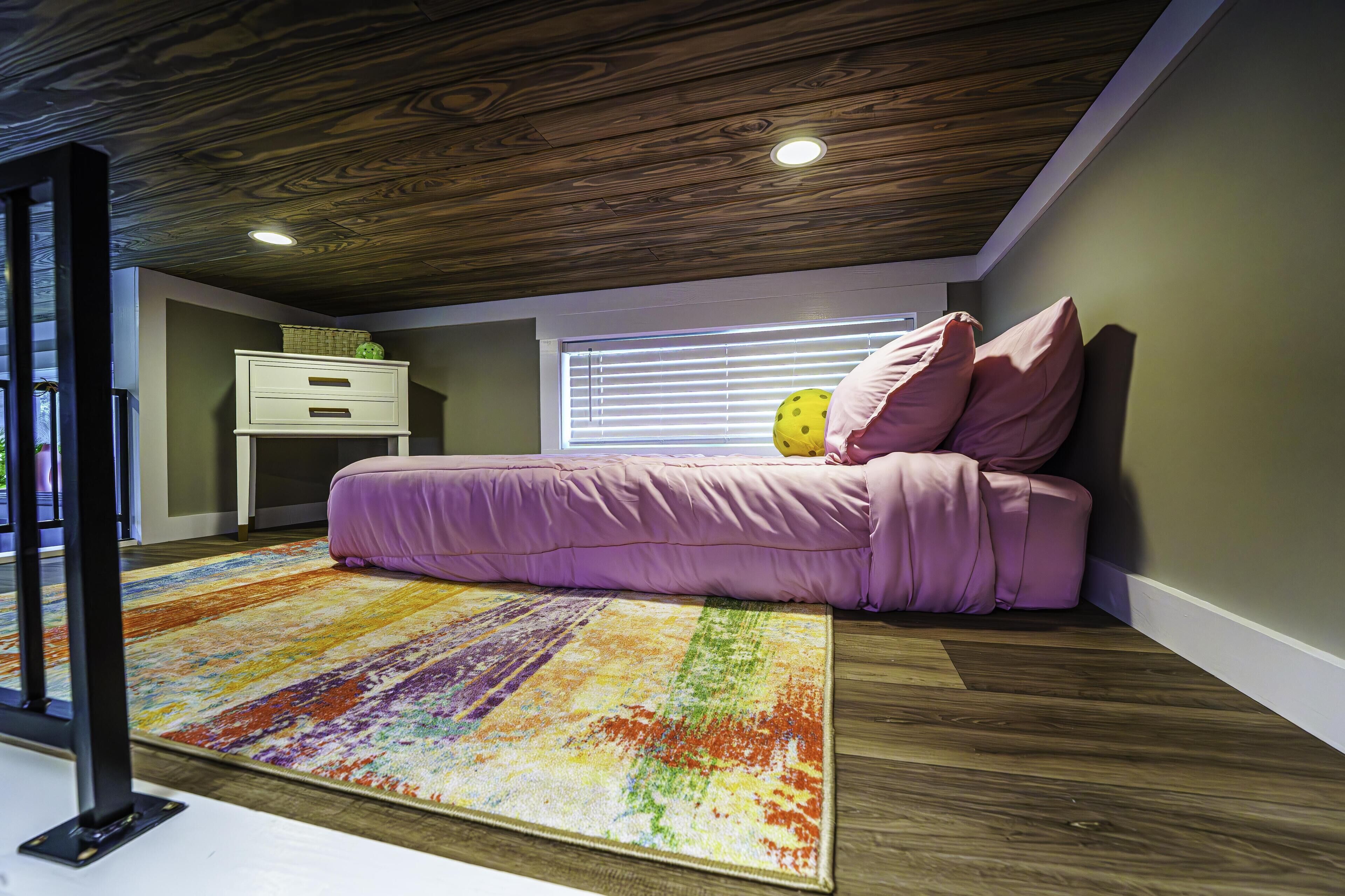 A bedroom with a wooden ceiling, a colorful rug, a bed with pink bedding, a nightstand, and a window with blinds.
