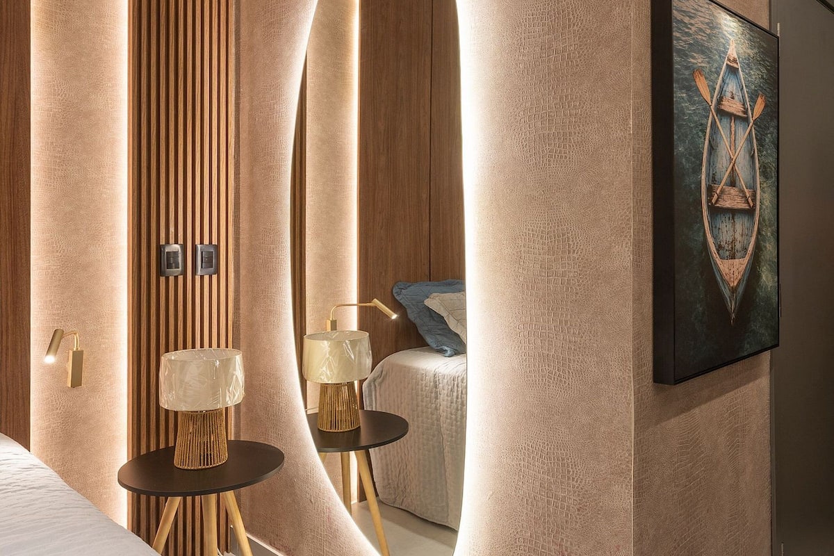A modern hotel room with a large mirror, a wooden panel wall, a small table with a lamp, and a framed artwork on the wall.