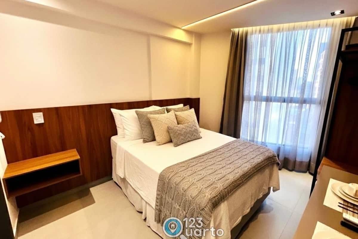A hotel room with a bed, a wooden headboard, a bedside table, a window with curtains, and a view of the city.