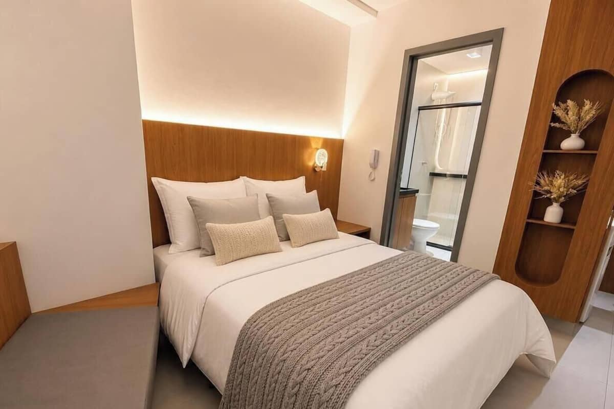 A modern hotel room with a large bed, a bedside table, a nightstand, a mirror, and a wooden shelf with decorative items.