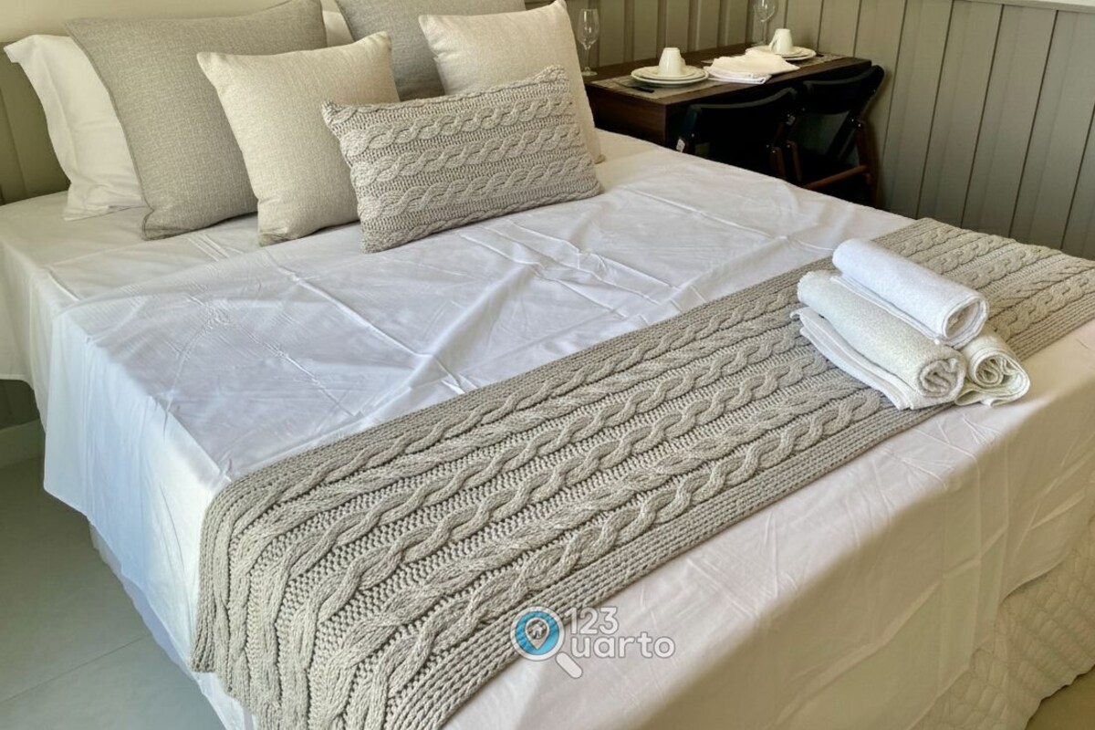 A neatly made bed with a white bedspread and a knitted throw blanket, a small table with a plate and a glass, and a chair.