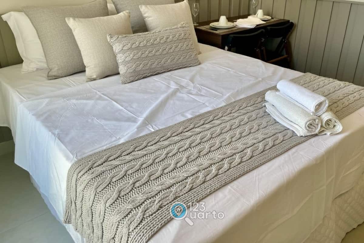 A neatly made bed with a white bedspread and a knitted throw blanket, a small table with a plate and a glass, and a chair.