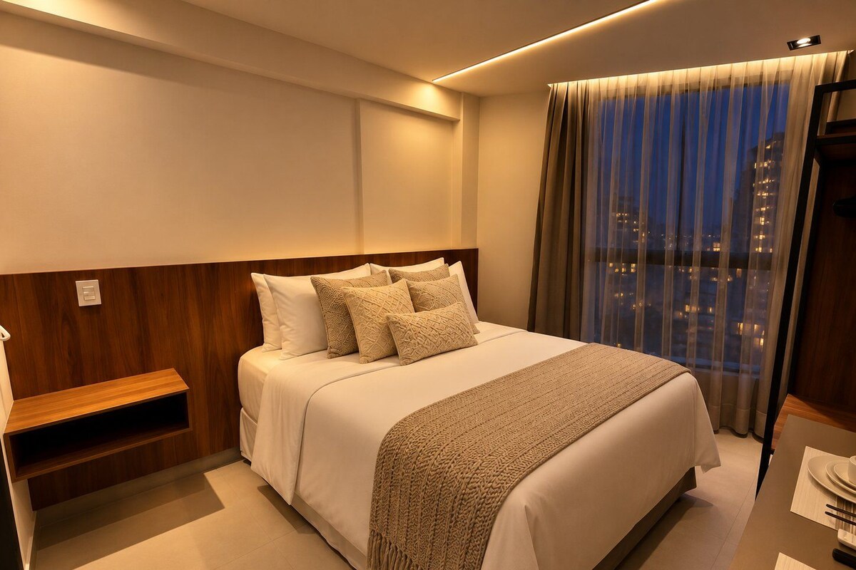 Quarto de hotel com uma cama grande, uma mesinha de cabeceira, vista da cidade à noite e uma janela com cortinas.