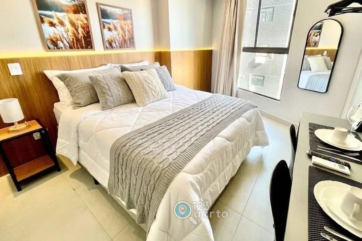 A hotel room with a bed, a bedside table with a lamp, a mirror, a window with curtains, and a view of the outside.