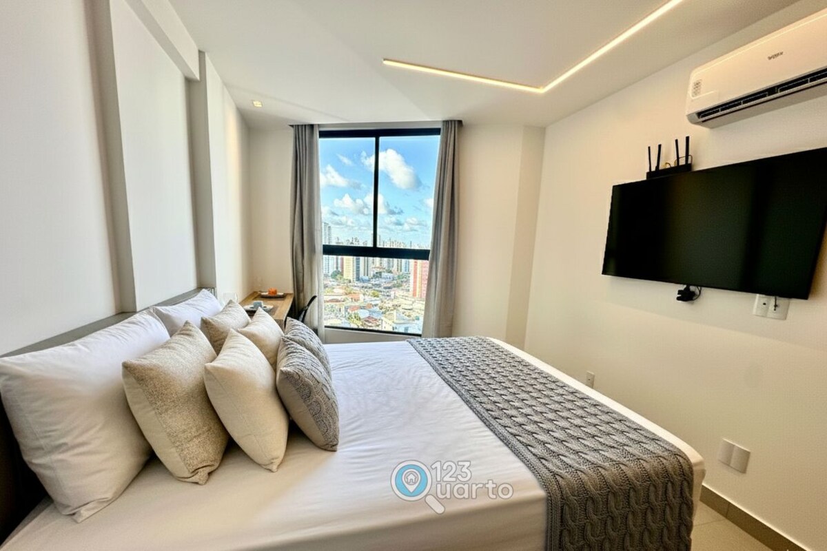 A hotel room with a bed, a TV, a window with a city view, and an air conditioning unit.