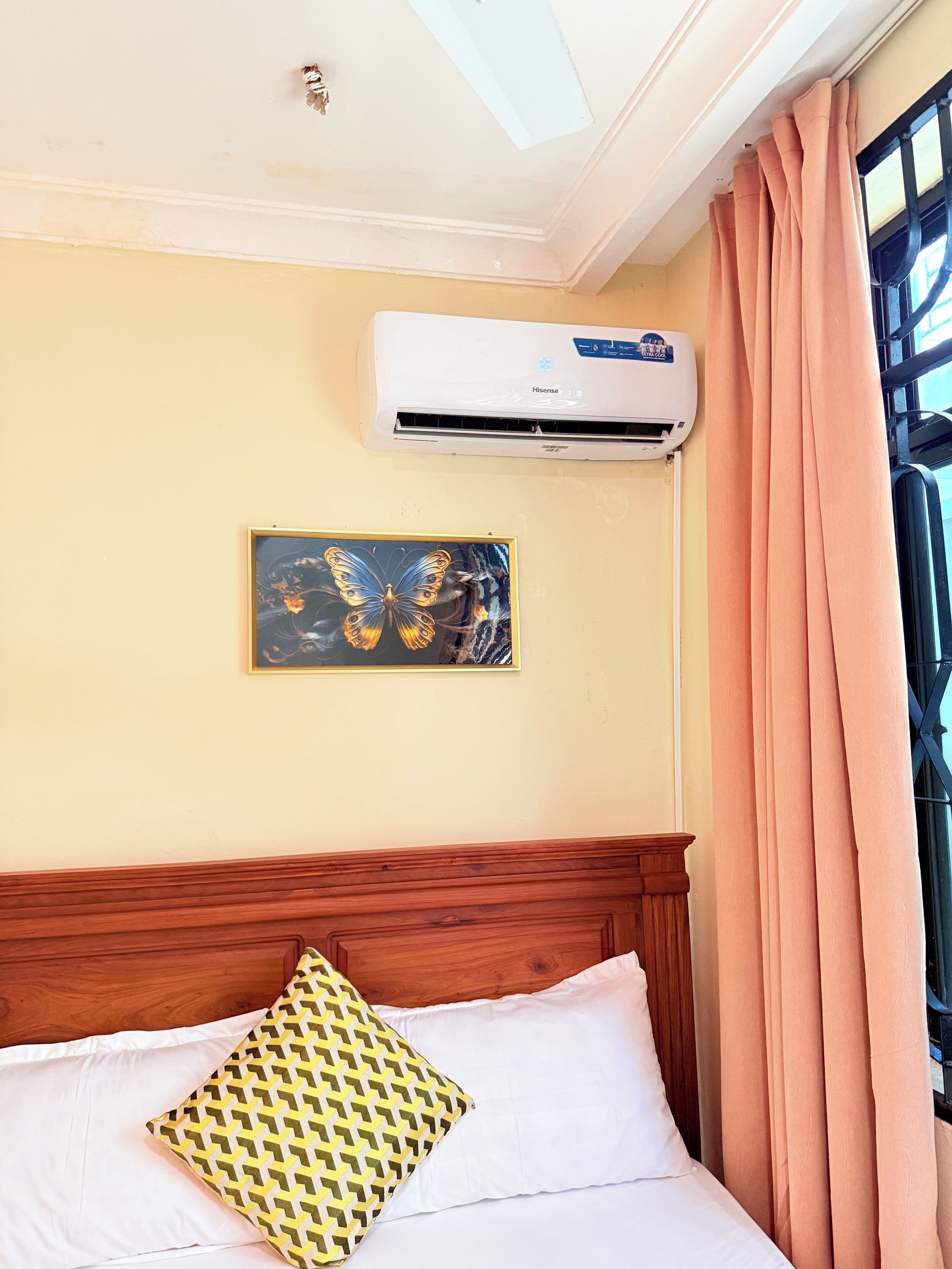 A hotel room with a bed, a wall-mounted air conditioner, a window with curtains, and a framed picture on the wall.