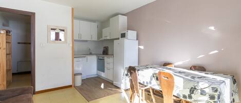 A compact kitchen with white cabinets, a microwave, and a refrigerator. There is a dining area with a table and chairs, and a wooden chair is visible.