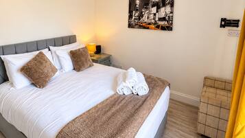 A neatly made bed with white linens and a brown throw blanket, a bedside table with a lamp, a wall-mounted power outlet, and a framed picture of a cityscape.